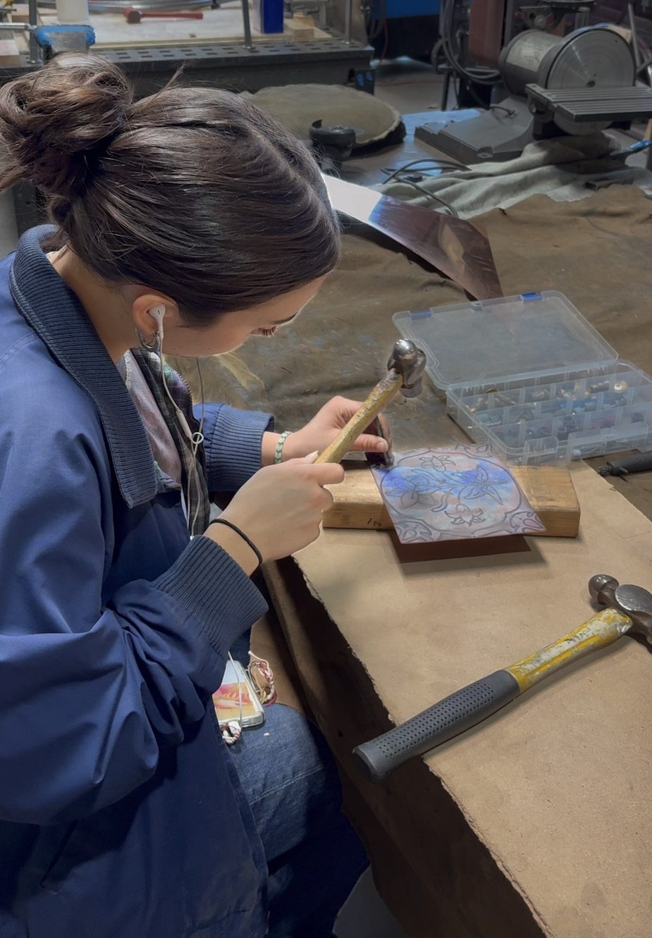 An apprentice using a hammer and punch to tool a floral repoussé design into a metal panel